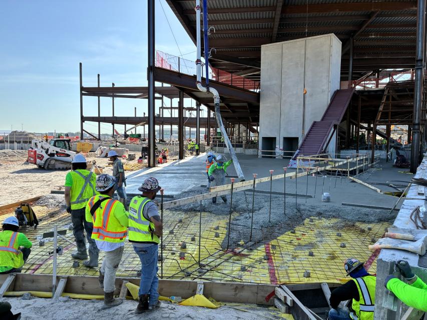 Construction workers pouring concrete from a supply tube into a form on the ground to create the flood of Airside D.